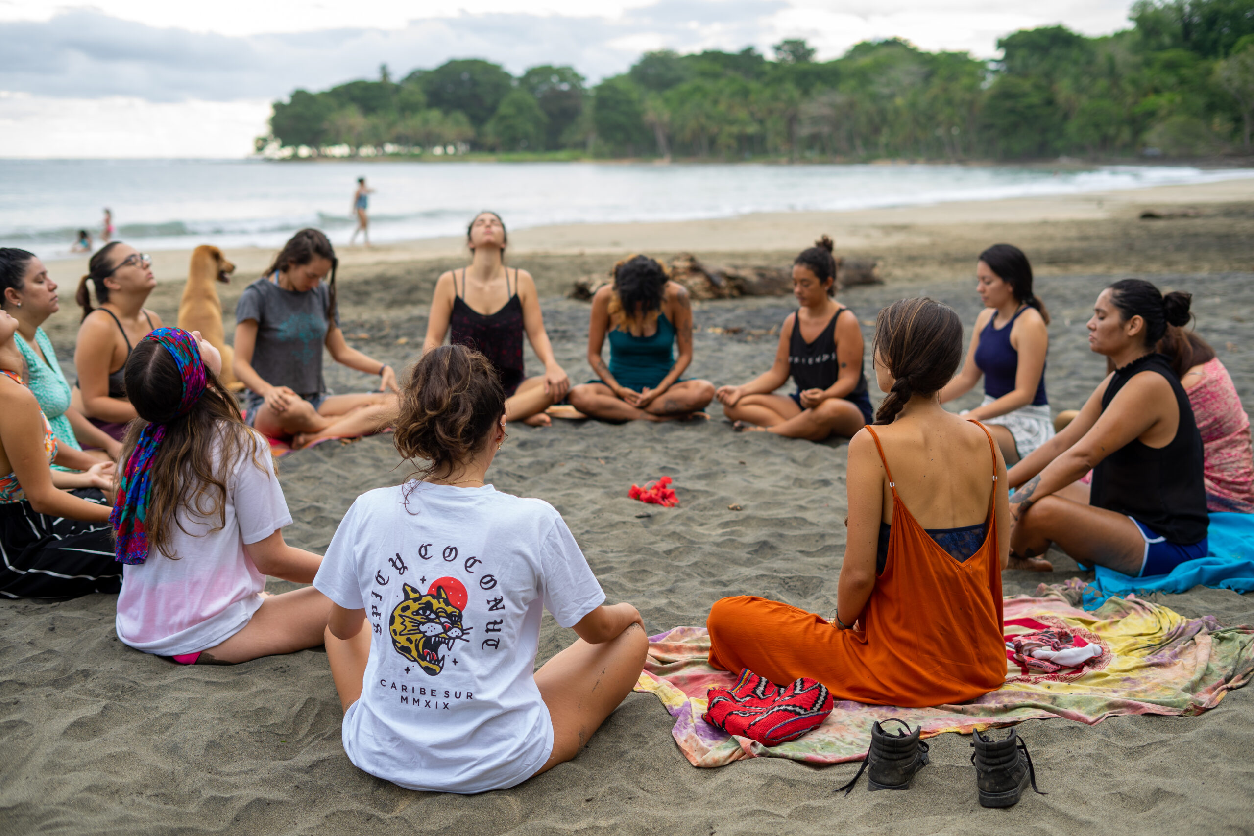 People gathered in a circle sitting cross-legged for a breathing exercise during a Toby Israel ESD self-defense workshop on the beach.